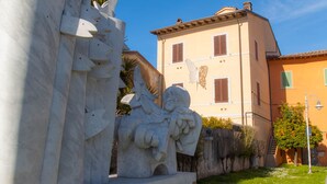 Une sculpture en pierre représentant une figure et un papillon, un bâtiment à la façade jaune et aux volets verts, et un ciel bleu clair.