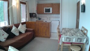 A compact living space with a brown sofa, a small wooden table with chairs, a kitchenette with a microwave and a kettle, and a window with curtains. - Apartment with three genuine bedrooms within 2 mins walk of the main lift (Morillon)