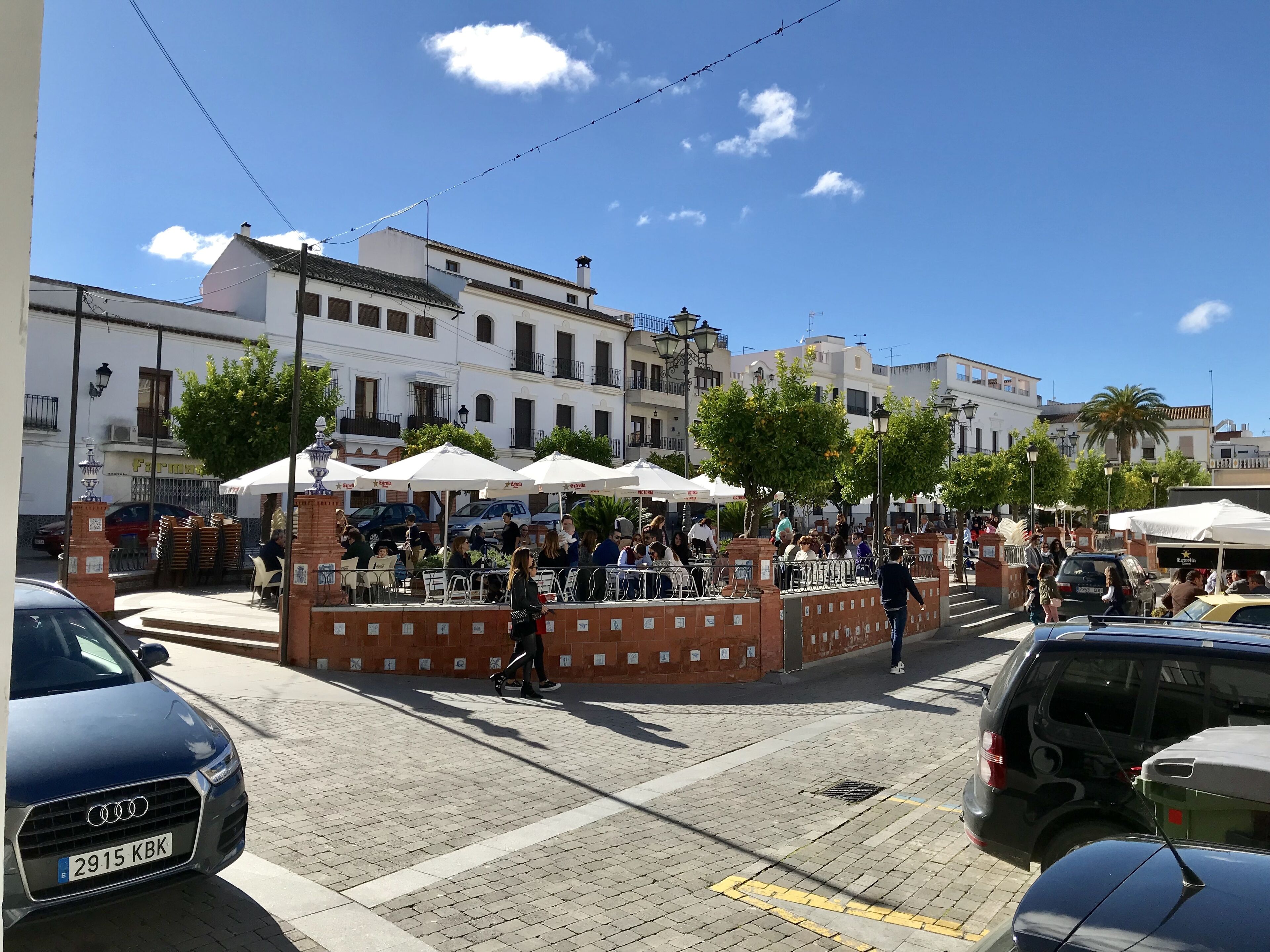 Una calle con terrazas al aire libre, coches aparcados a un lado y edificios al fondo.