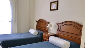 A hotel room with two beds, each with a blue bedspread and white pillows. There is a wooden headboard on each bed, a nightstand with a lamp, and a framed picture on the wall.
