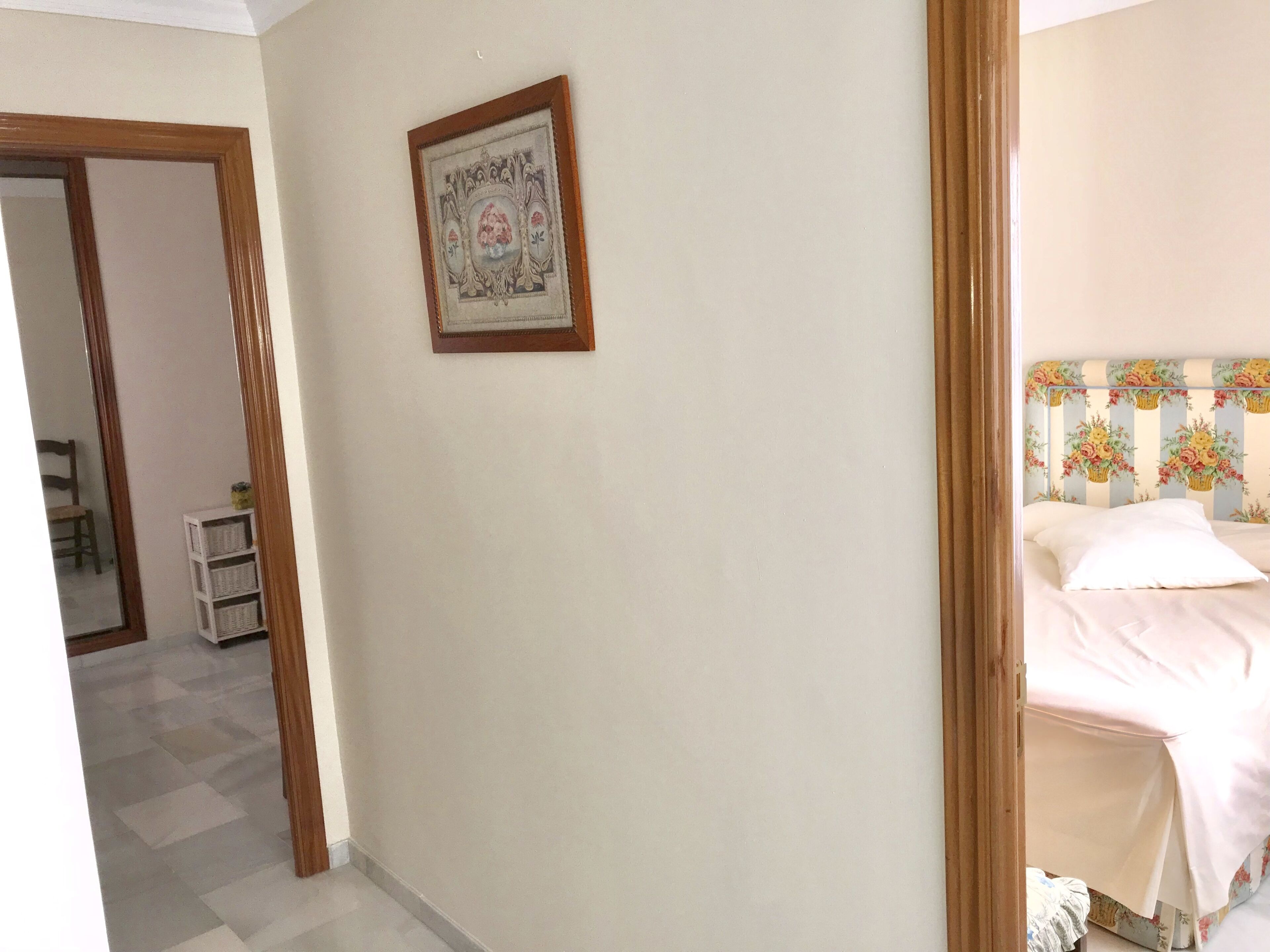 Un dormitorio con cama, mesita de noche, cuadro enmarcado en la pared y puerta que da a otra habitación.