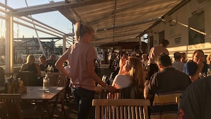 An outdoor dining area with wooden tables and chairs, a covered roof, and a view of the marina in the background.