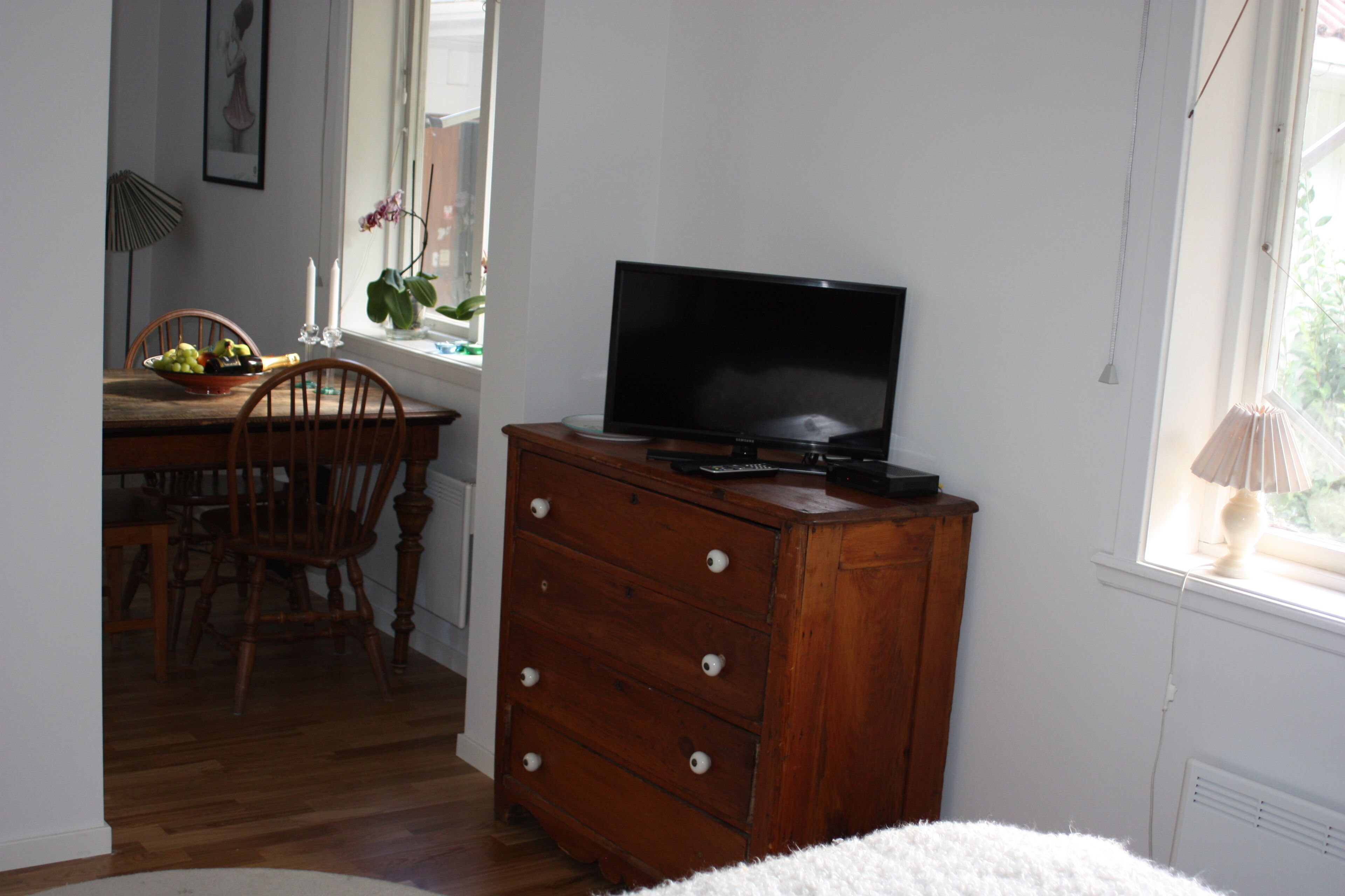 A room with a wooden dresser, a television on top, a dining table with chairs, and a window with a plant.