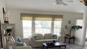 A living room with a sofa, coffee table, and a lamp. There is a ceiling fan and a window with blinds.