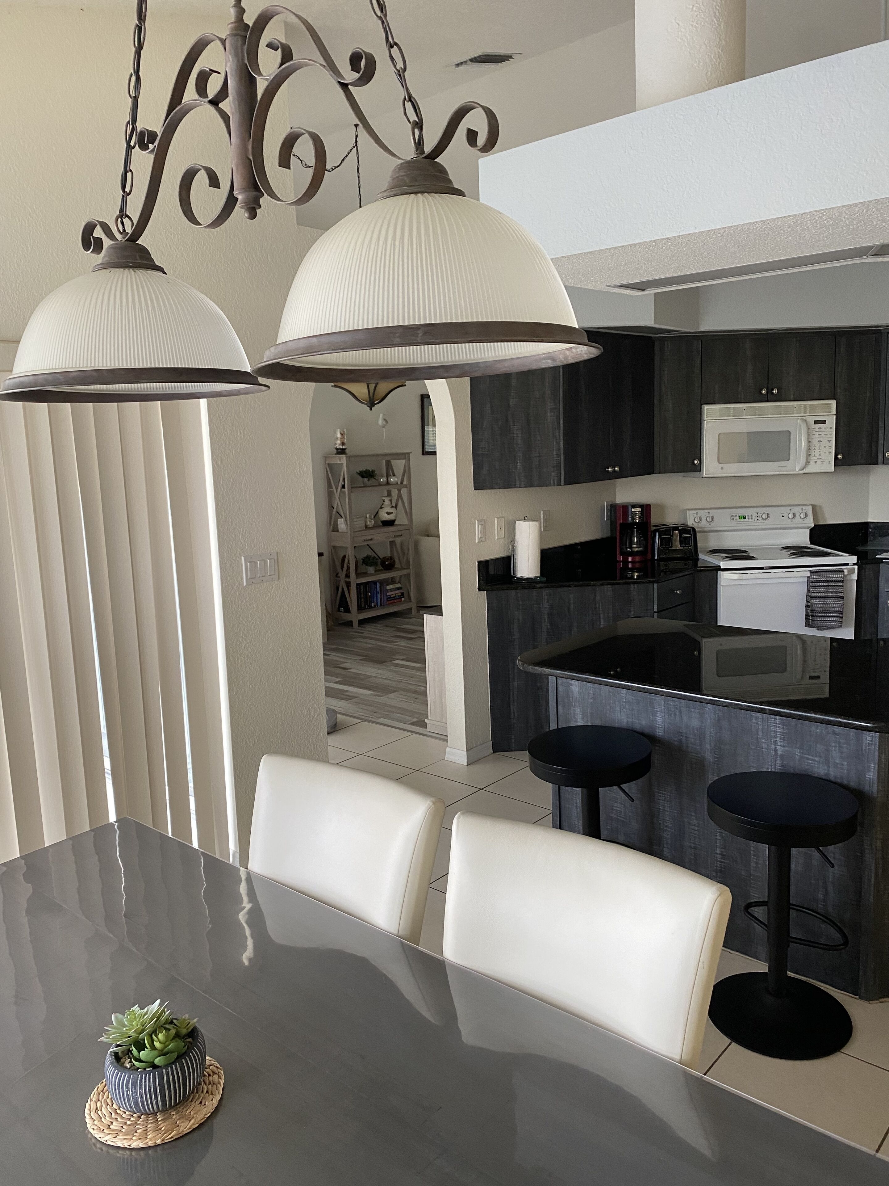 Una cocina moderna con una isla central, armarios blancos y un microondas. Hay una zona de comedor con una mesa y sillas, y un candelabro colgando de arriba.