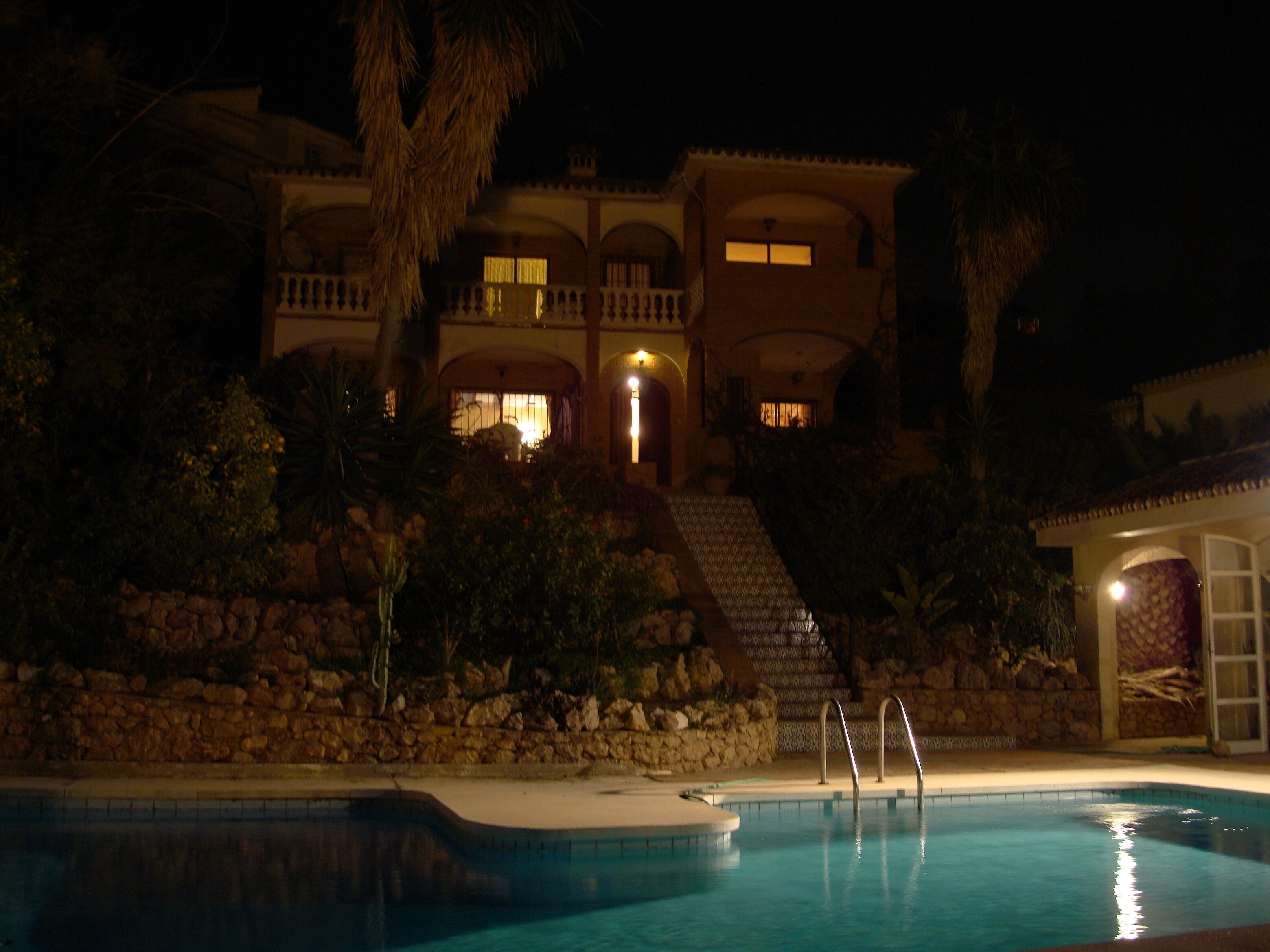 Vue nocturne d’un complexe hôtelier avec une piscine, un mur de soutènement en pierre et un immeuble à plusieurs étages dont les fenêtres sont illuminées.