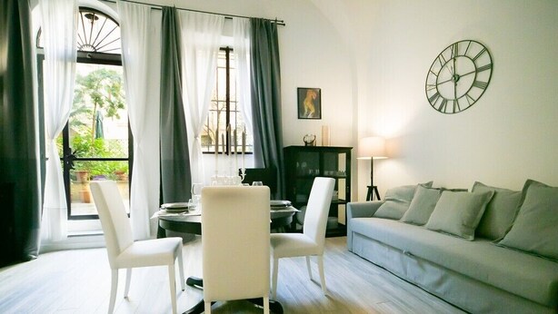 A living room with a grey sofa, white chairs, a dining table, and a clock on the wall.