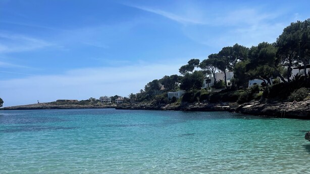 A beach with clear turquoise water, sandy shore, and buildings with trees in the background. - Beachfront design villa in Bauhaus style, pool, highspeed internet, air conditioning / heating (Cala Murada)