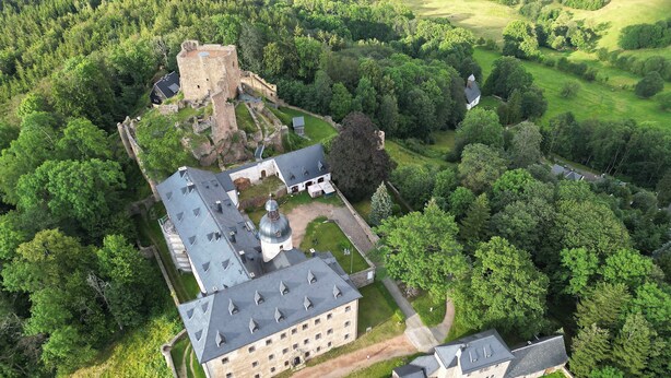 An aerial view of a castle complex with a central building featuring a large dome, surrounded by lush greenery and a moat.