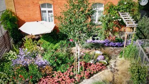A garden with a variety of plants and flowers, a wooden pergola, and a brick building with windows in the background.
