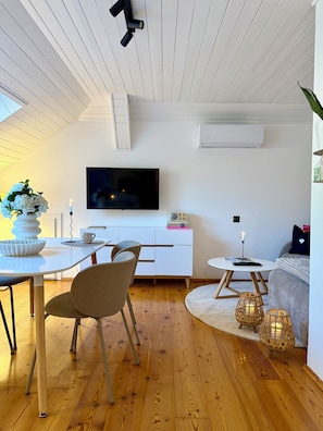 Un soggiorno moderno con televisore a schermo piatto, arredi bianchi, pavimento in legno e soffitto a vista con travi.