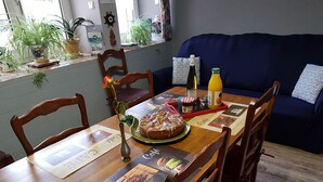 A dining table with a cake, a vase with a flower, a bottle of orange juice, and a bottle of wine. There are chairs around the table, and a blue couch in the background.