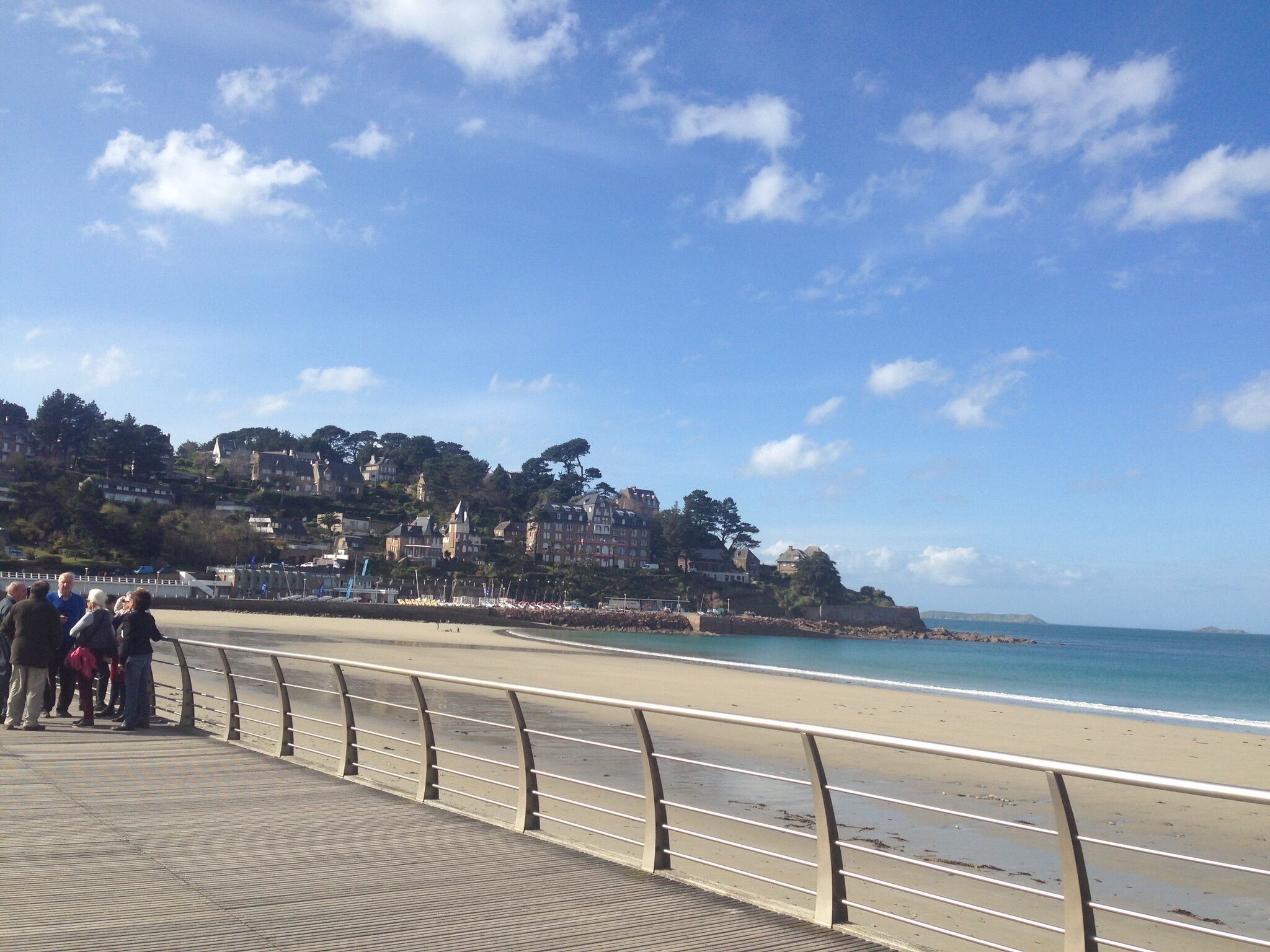 A coastal promenade with a metal railing, a sandy beach, and a backdrop of buildings on a hillside.