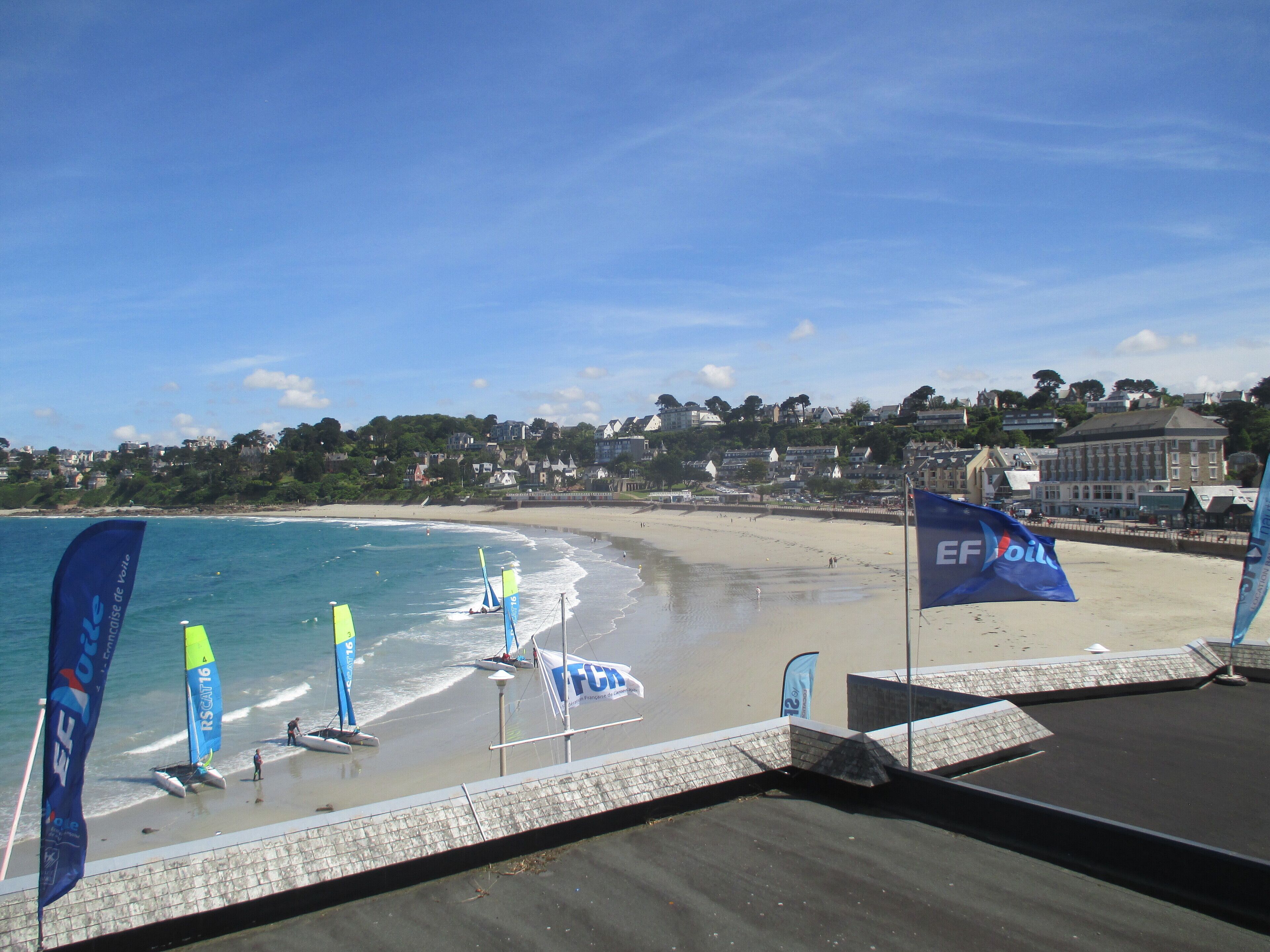 A beach with flags, a sandy shore, and buildings in the background.