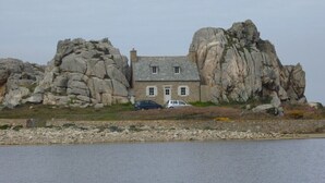 A house with a stone exterior, situated between two large rock formations, overlooking a body of water. - Recent south-facing apartment 2 steps from the beach with private parking (Perros-Guirec)