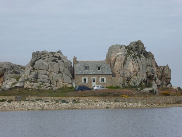 A house with a stone exterior, situated between two large rock formations, overlooking a body of water.