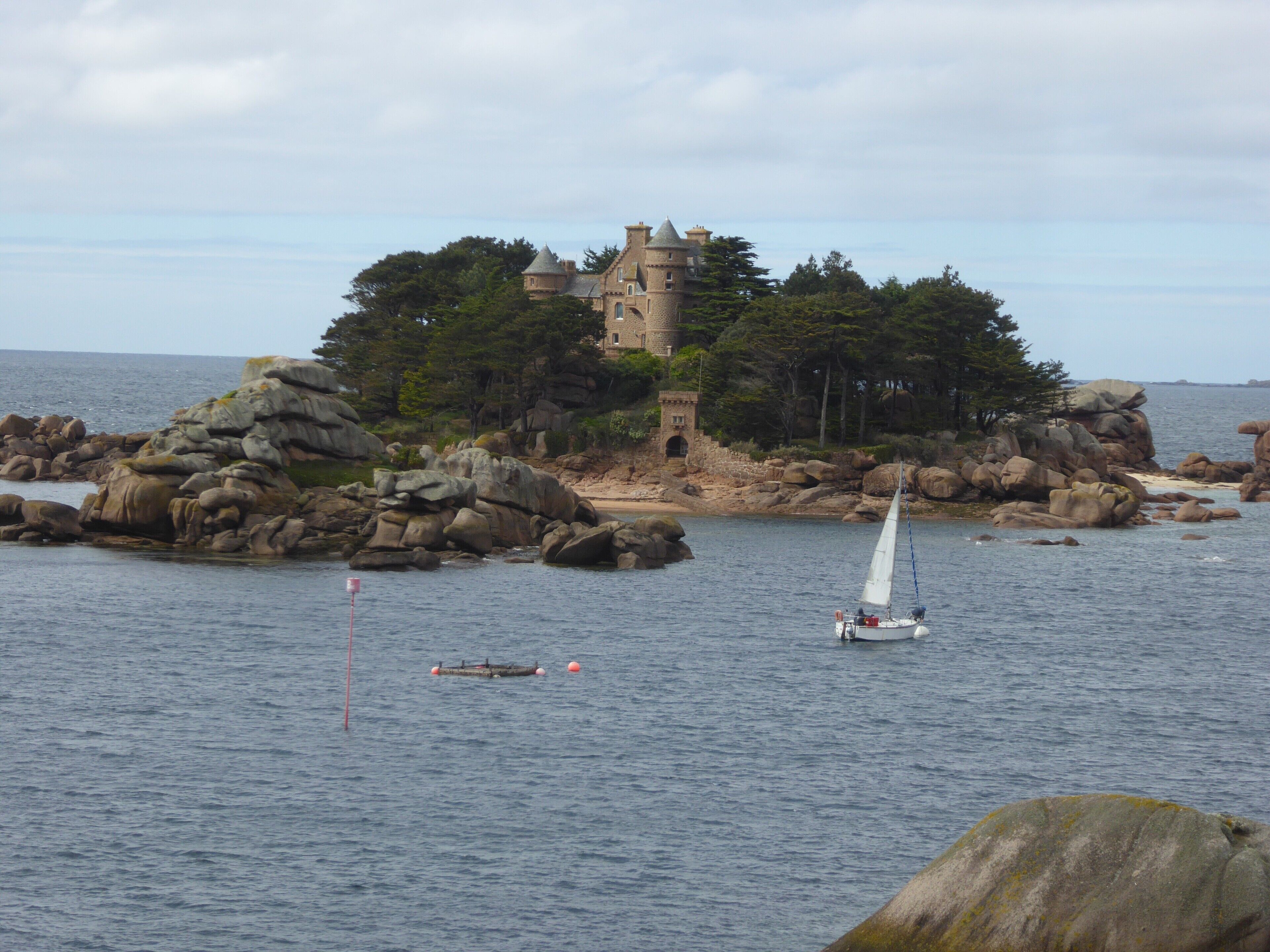 A coastal scene with a castle on a rocky island, a sailboat on the water, and a buoy.