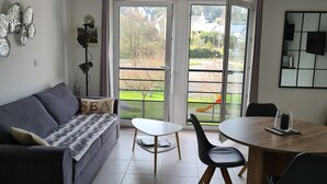 A modern living room with a sofa, a coffee table, and a dining table with chairs. There are large windows with a view of the outside. - Recent south-facing apartment 2 steps from the beach with private parking (Perros-Guirec)