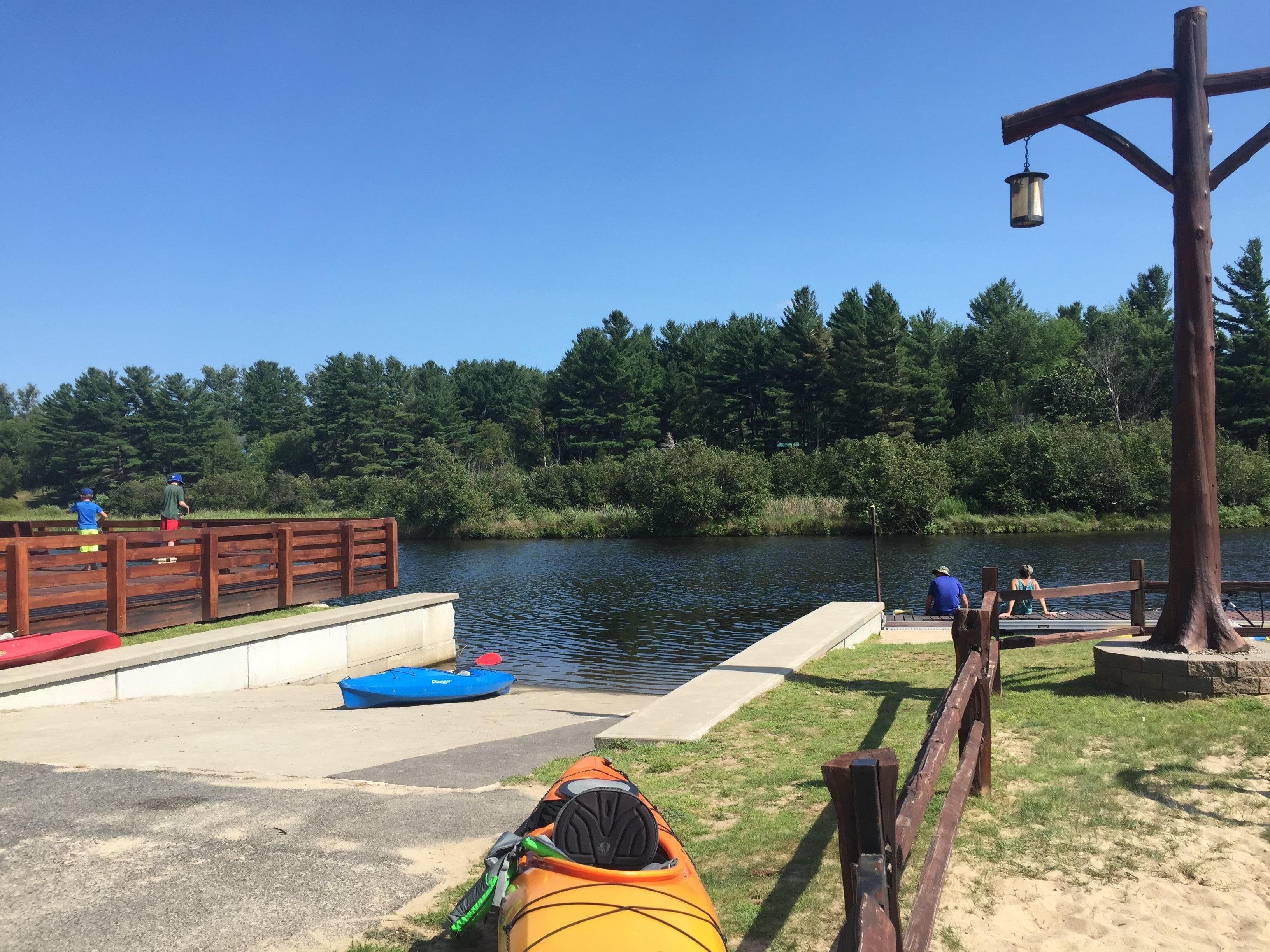 Un muelle de madera con kayaks y un bote de remo, una plataforma de concreto y una orilla fluvial arbolada.