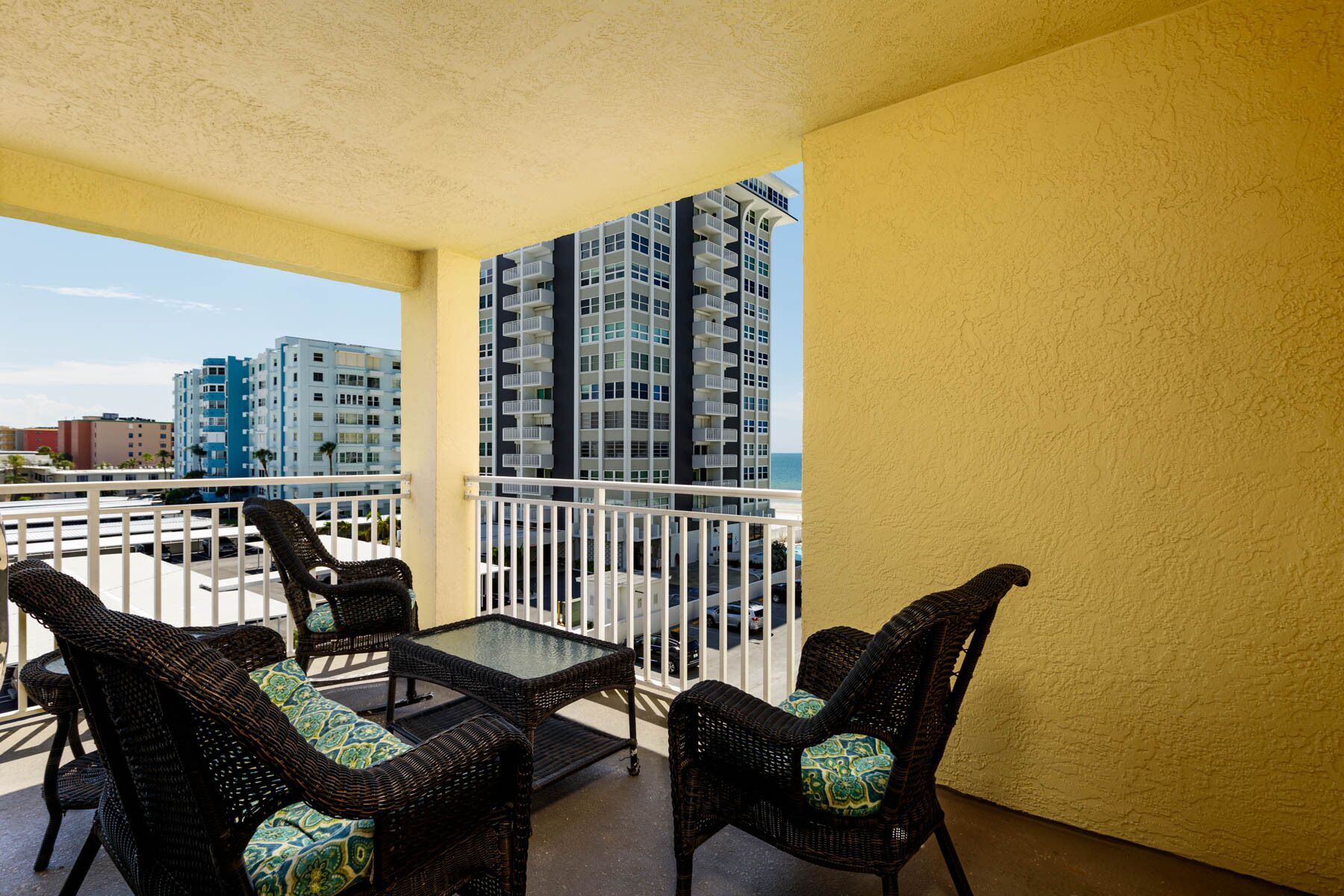 A balcony with wicker furniture and a glass table, overlooking a high-rise building and a parking area.