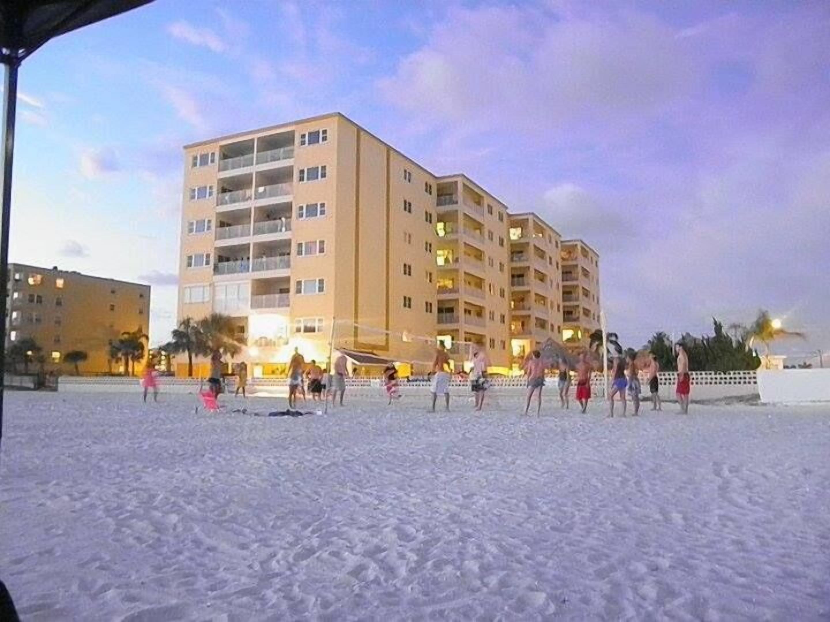 Un hotel fronte spiaggia con diversi piani e balconi, una spiaggia sabbiosa in primo piano e palme sullo sfondo.