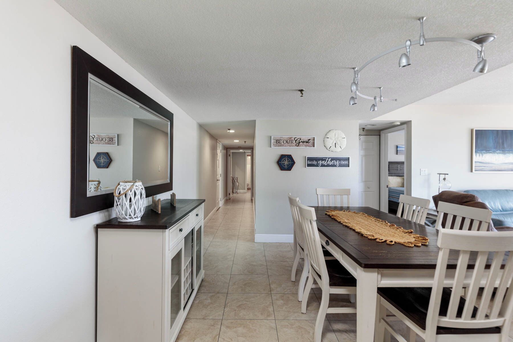 Una cocina moderna con encimera oscura, armarios blancos y un espejo grande. Hay una zona de comedor con mesa de madera y sillas blancas, y un pasillo que conduce a otras habitaciones.