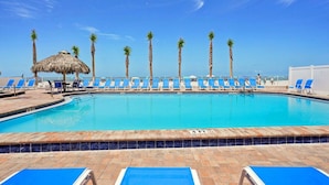A swimming pool with blue lounge chairs, palm trees, and a thatched umbrella.
