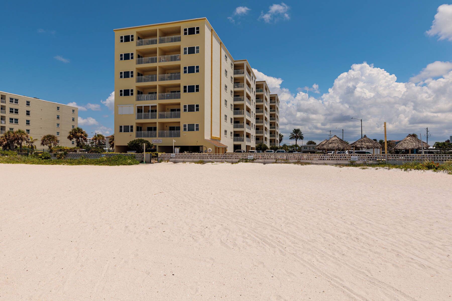 Un hôtel à plusieurs étages doté de balcons, situé près d’une plage de sable et bordé de constructions aux toits de chaume.