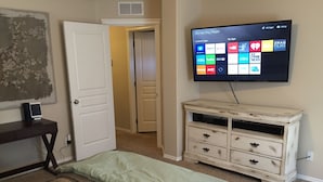 Un dormitorio con cama, cómoda, televisor montado en la pared mostrando diversas aplicaciones, y una puerta que da a otra habitación.