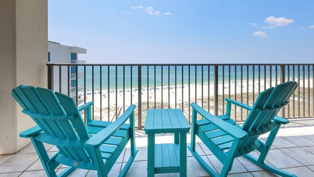 A balcony with turquoise Adirondack chairs and a small table, overlooking a beach with a clear view of the ocean and a building in the background.