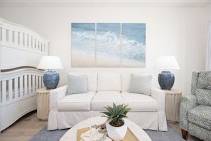 Un salon avec un canapé blanc, deux tables d’appoint avec des lampes, une grande peinture murale représentant des vagues et une plante en pot posée sur une table basse.