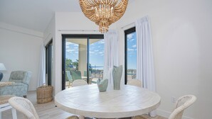 A round dining table with wicker chairs, a chandelier, and a view through large windows.