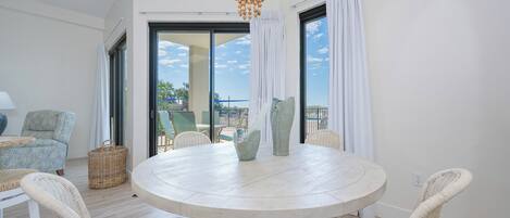 A round dining table with wicker chairs, a chandelier, and a view through large windows.