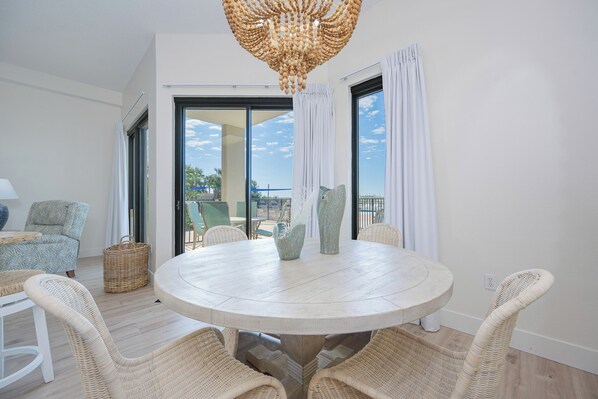 A round dining table with wicker chairs, a chandelier, and a view through large windows.