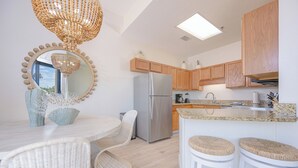 A modern kitchen with wooden cabinets, a stainless steel refrigerator, a dining area with wicker chairs, and a bar with stools.