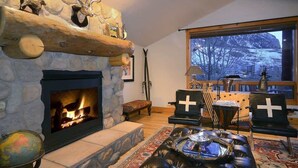 Une cheminée en pierre avec un feu de bois, un pouf en cuir, un globe terrestre et une grande fenêtre donnant sur les arbres.