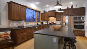 Une cuisine moderne avec des meubles en bois, un grand îlot central, des appareils électroménagers en acier inoxydable et une fenêtre donnant sur un arbre.