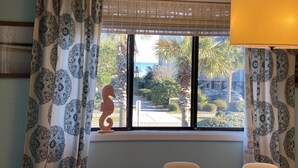 A room with patterned curtains, a wooden seahorse decoration on the windowsill, and a view of a street and palm trees outside. - Modern, Completely Renovated 2 bdrm 2 bath Ocean View (Surfside Beach)