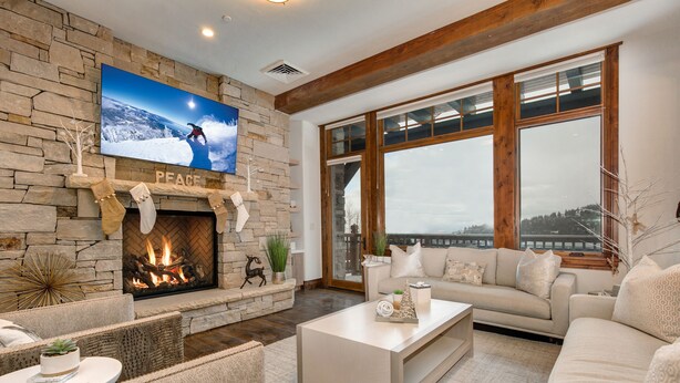 A cozy living room with a stone fireplace, a large TV mounted above it, and a view of the outdoors through large windows.