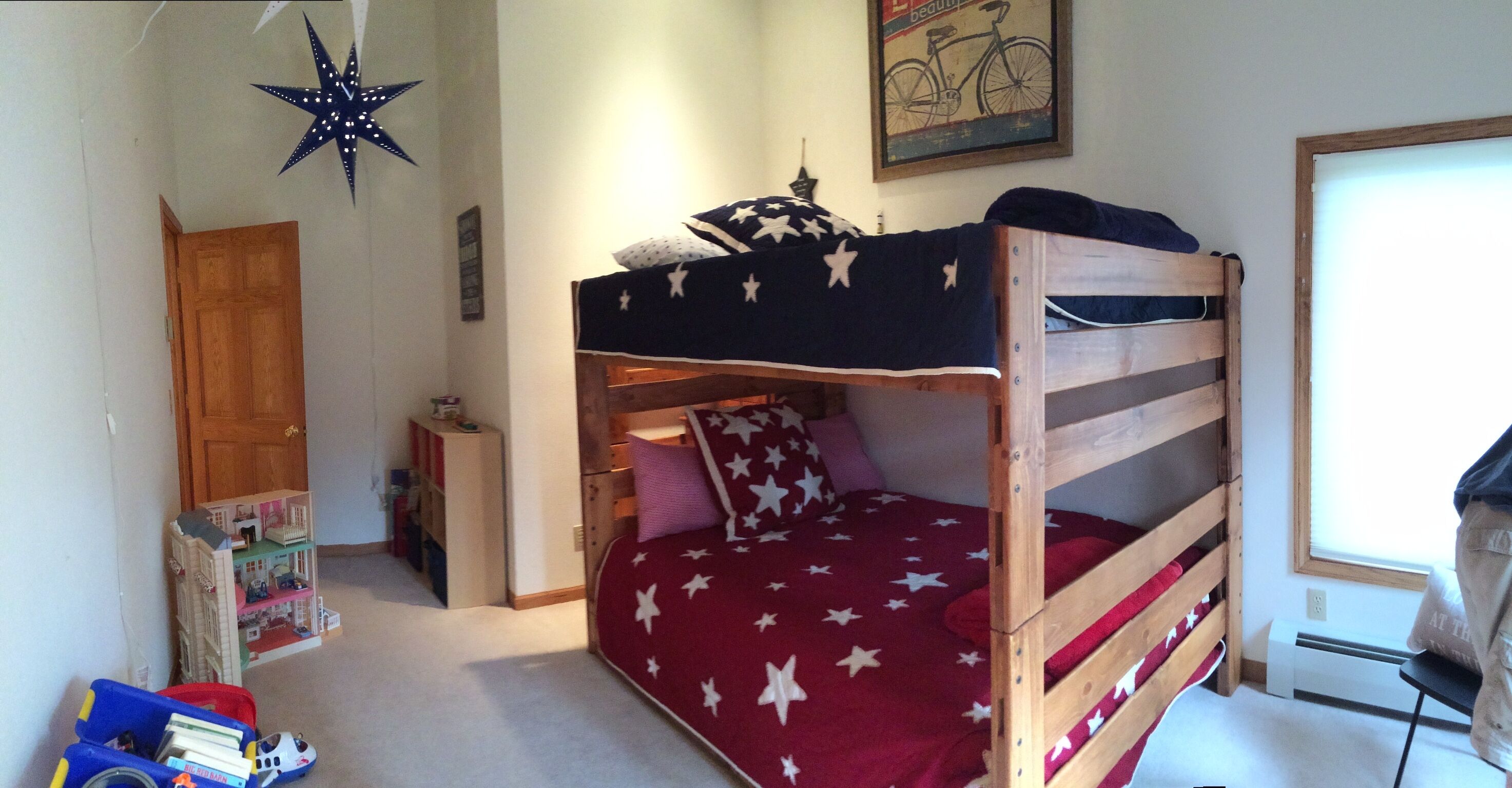Un lit superposé avec une couverture à motifs d’étoiles, une porte en bois, une petite table avec une lampe et une affiche de vélo accrochée au mur.