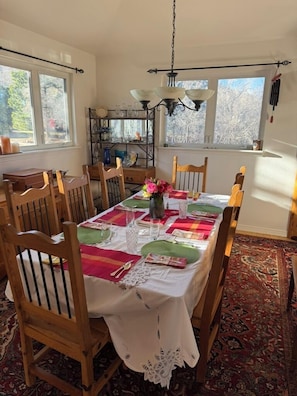 A dining room with a table set for six, surrounded by wooden chairs, a patterned rug, and a decorative shelf with glassware.