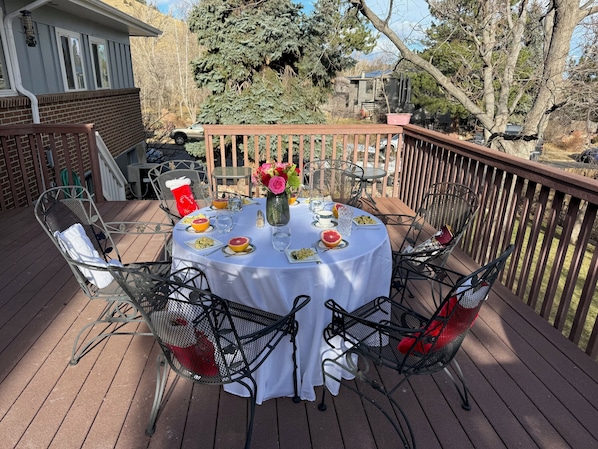 A round table set for four on a wooden deck with metal chairs, a white tablecloth, and a vase of flowers.