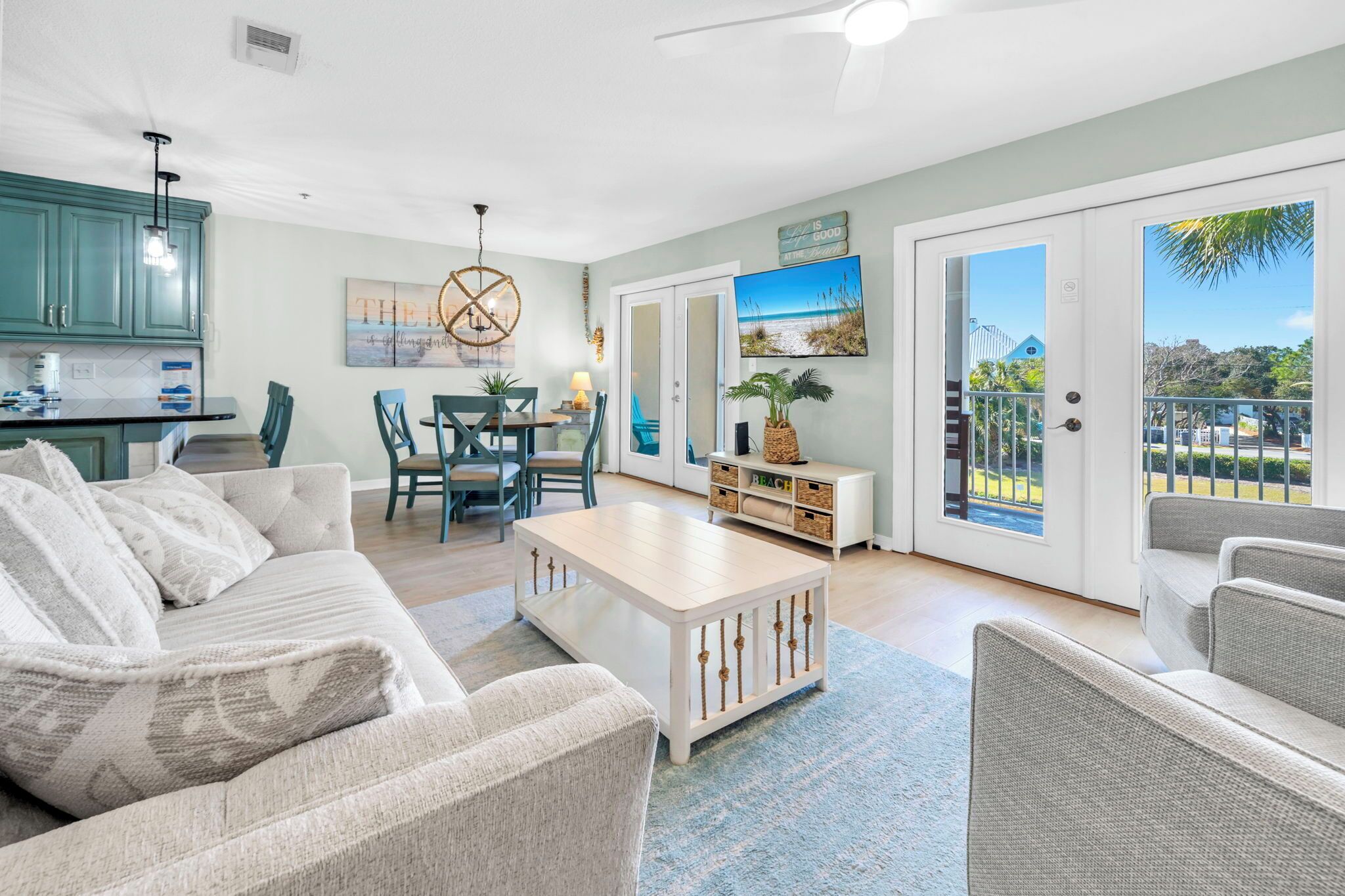 A spacious living area with a sofa, coffee table, and a dining table with chairs. There is a kitchen area with teal cabinets and a pendant light. A large window offers a view of the outdoors.