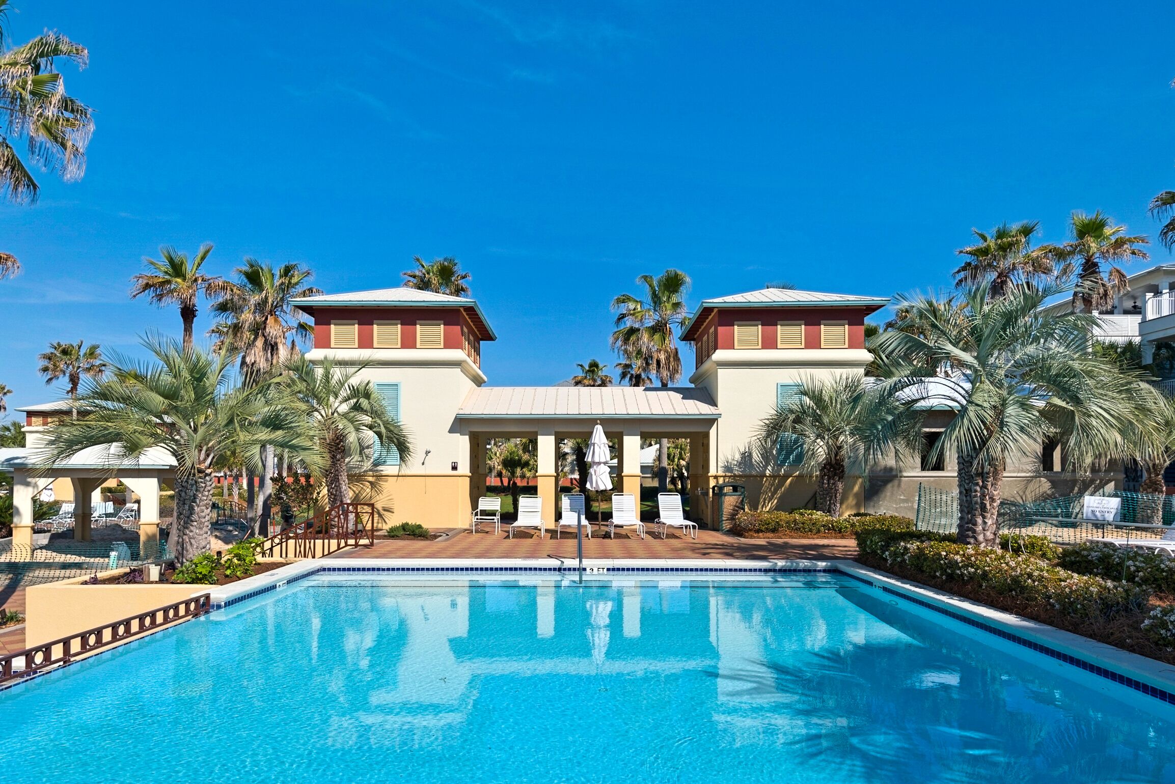 Una piscina con sedie a sdraio e palme, circondata da edifici con una combinazione di colori rosso e beige.