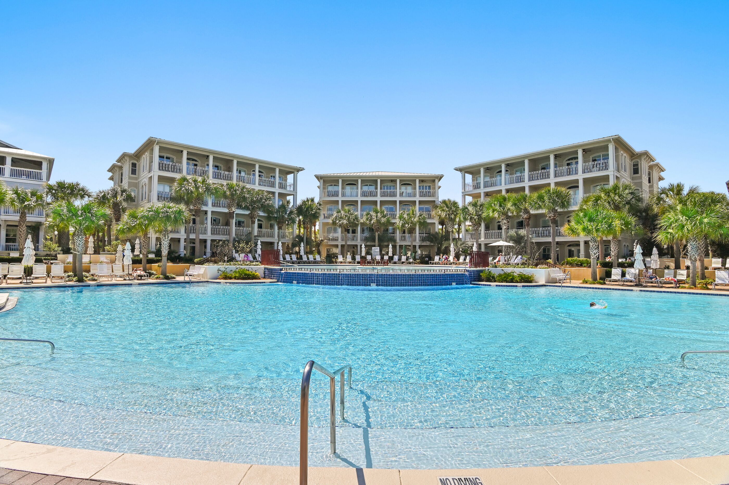 A swimming pool with a blue deck, surrounded by palm trees and lounge chairs, with a building featuring balconies in the background.