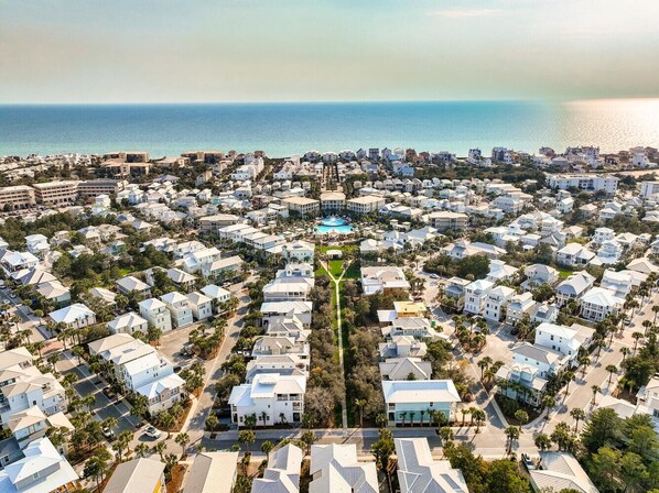 An aerial view of a coastal residential area with houses, a central road, and a body of water in the background.