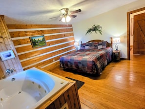 A bedroom with a wooden slat wall, a large bed with a patterned quilt, a wooden floor, a ceiling fan, and a bathroom with a bathtub.