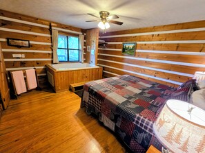 Ein Blockhauszimmer mit Holzvertäfelung, einem großen Bett, einem fenster mit Vorhängen, einem Deckenventilator und einer Holzbank.