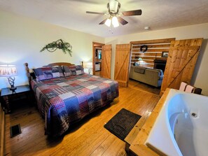 A bedroom with a bed, a wooden floor, a ceiling fan, a nightstand, a lamp, a wall-mounted plant, and a bathroom with a bathtub. - B's Nest a Private & Clean w Mtn Views & Hot Tubs! (Sevierville)
