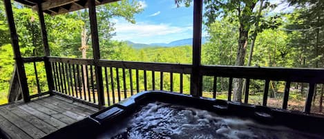 A hot tub with a wooden deck and a view of a forest and mountains.
